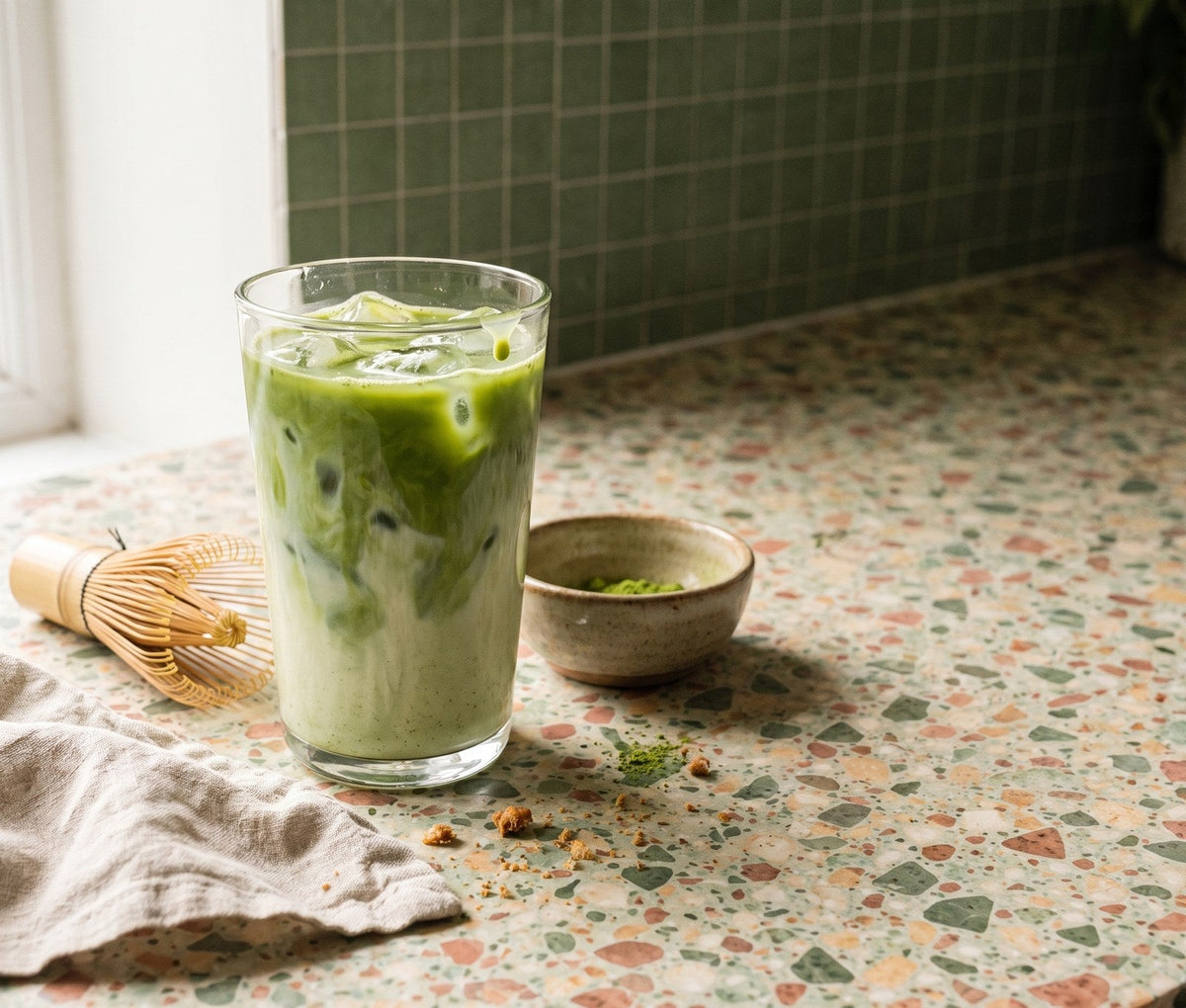 An iced matcha latte in a tall glass with a bamboo whisk and a little bowl of matcha
