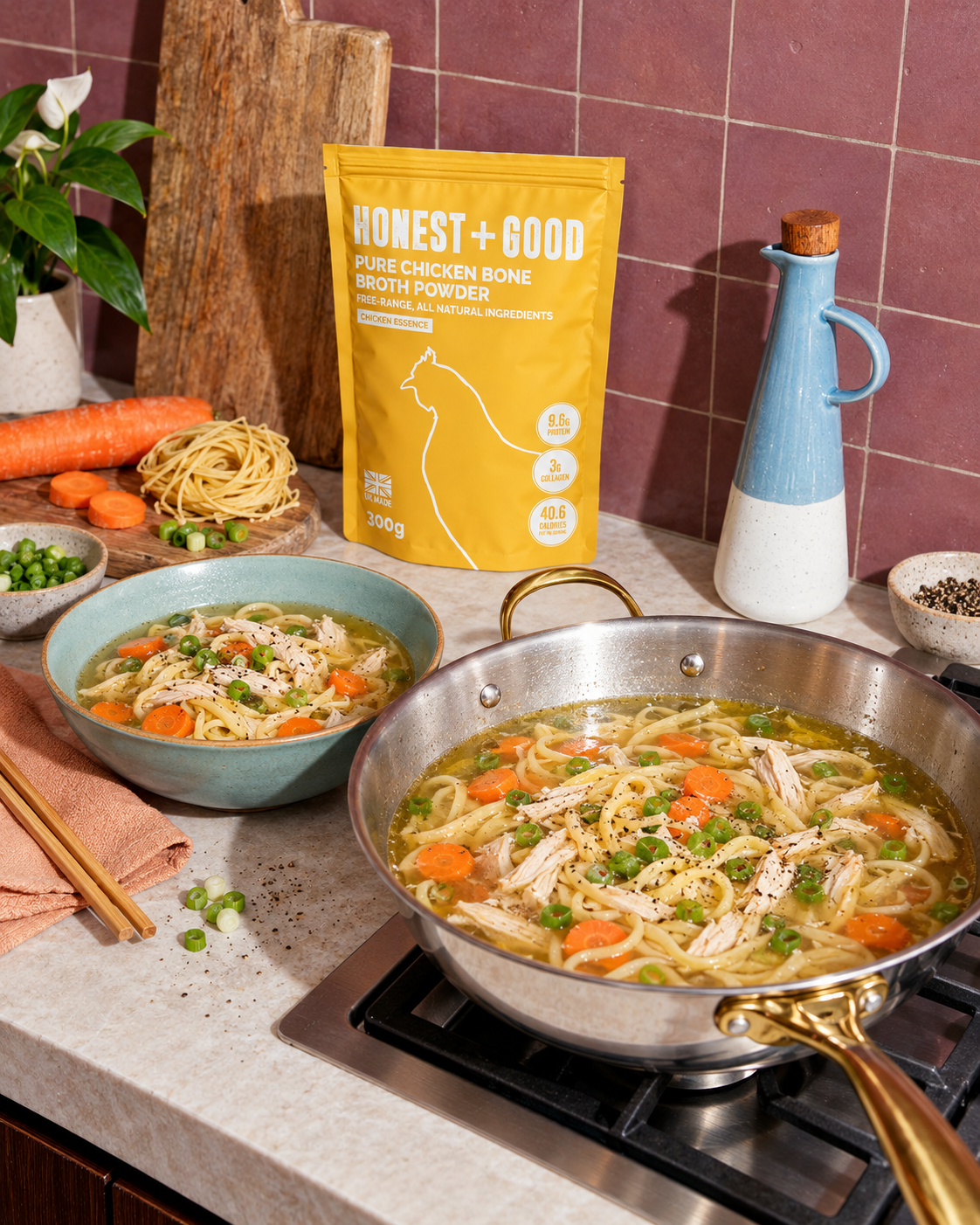 A yellow bag of Honest + Good Pure Chicken Bone Broth Powder stands on a kitchen counter beside a stainless steel pan of prepared chicken noodle soup with carrots and peas, with bowls and cooking props nearby.