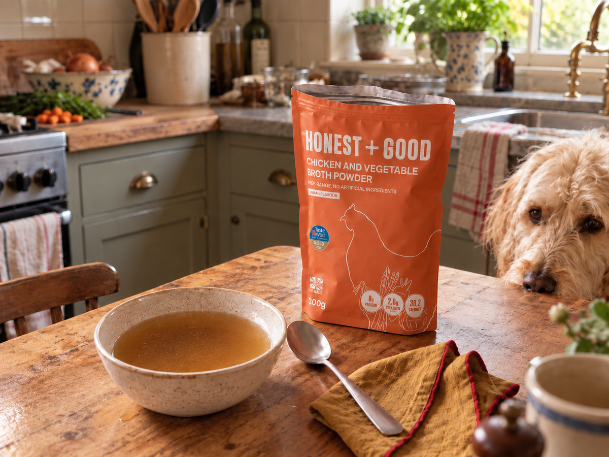 A bowl of broth on a kitchen table next to an Honest + Good Chicken + Vegetable pouch, with a hopeful dog peeking over
