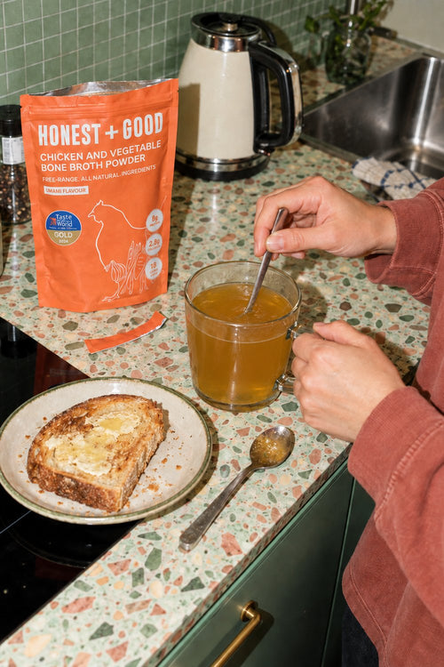A person stirs a glass mug of golden chicken and vegetable bone broth powder in a kitchen setting, with a plate of toast and an orange bag of Honest + Good bone broth powder on the countertop.